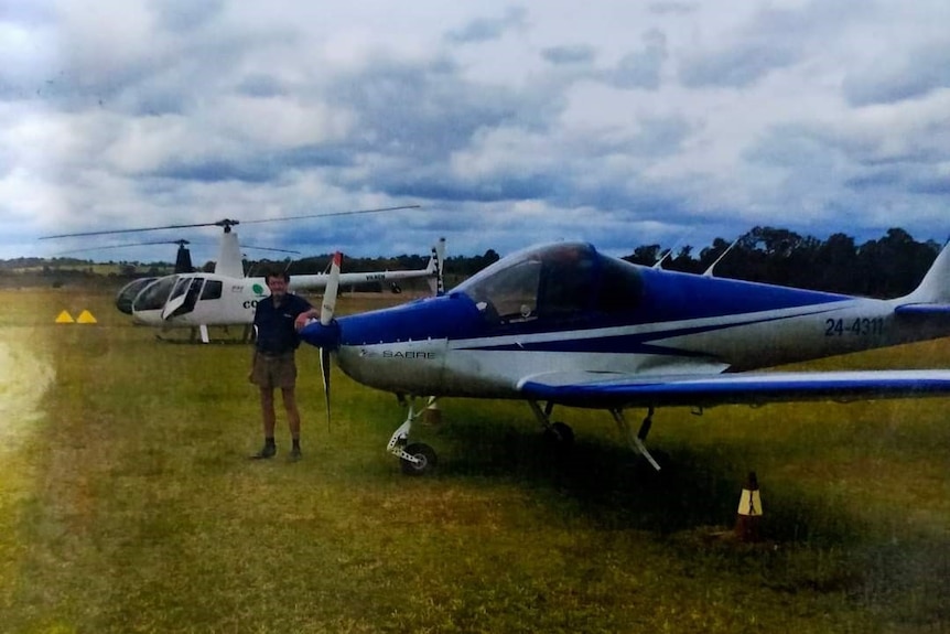 A man stands at the front of a small aeroplane. There is a small helicopter in the background.