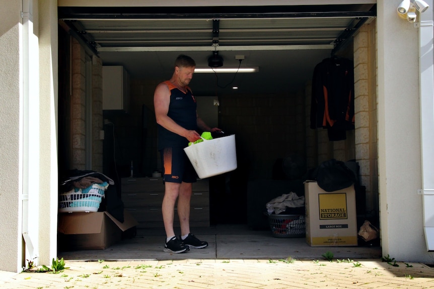 A man wearing a sleeveless shirt, shorts and joggers stands at the entrance to a garage carrying a laundry basket.