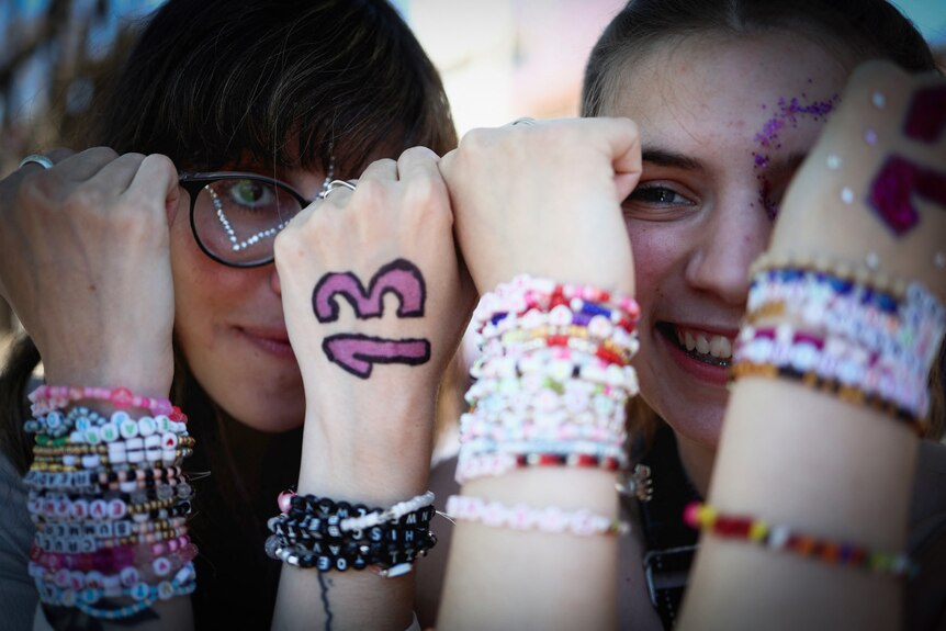 Two young women hold up their friendship bracelets and hands with the number 13 written on it.