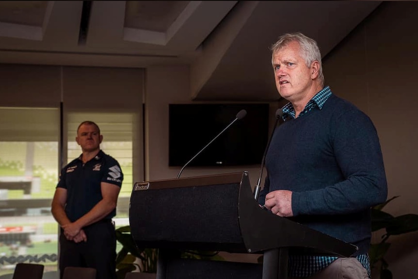 Man in blue check shirt and blue jumper stands at mic delivering speech.