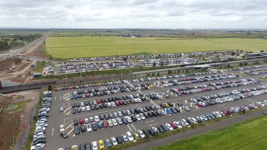 A drone shot of a large car park, filled with cars, near a train station and some green fields.
