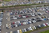 A drone shot of a large car park, filled with cars, near a train station and some green fields.