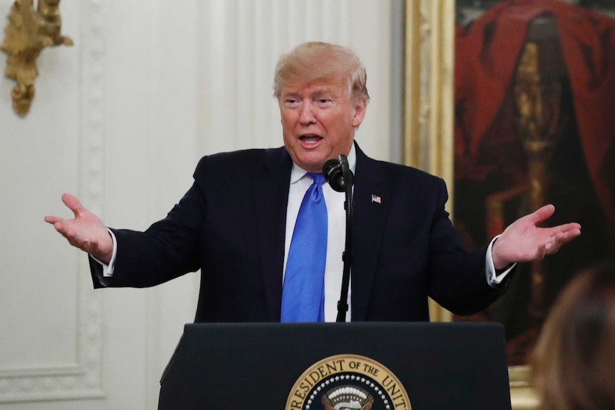 Donald Trump stands at a lectern with his hands up in a shrugging motion