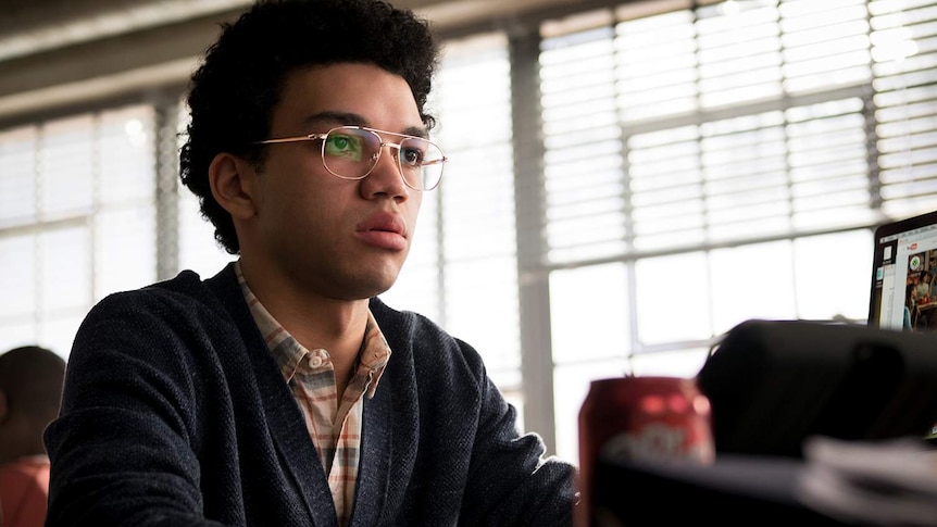 A young man with aviator spectacles looks worriedly at the scene in front of him.