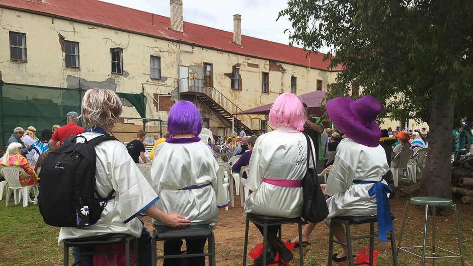 The backs of four people with colourful wigs sitting on stools with an old hotel facade in the background