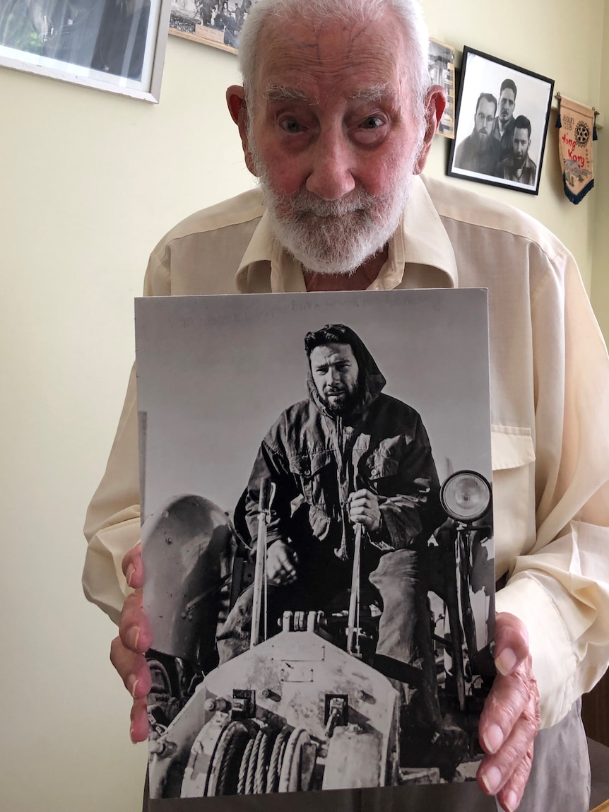 An elderly man holds a photo of himself from 60 years earlier, sitting on a tractor in Antarctica.