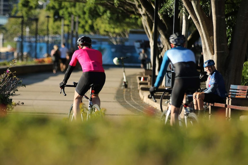 Two cyclists on a riverside path as another cyclist on a bench watches on