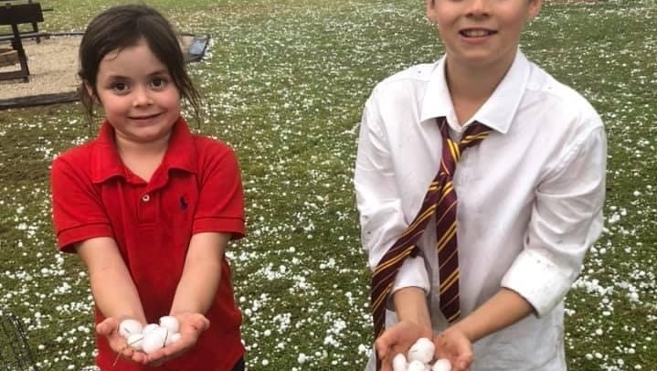A young girl and boy hold hail stones in their outreached hands smiling at the camera. The ground littered in hail stones