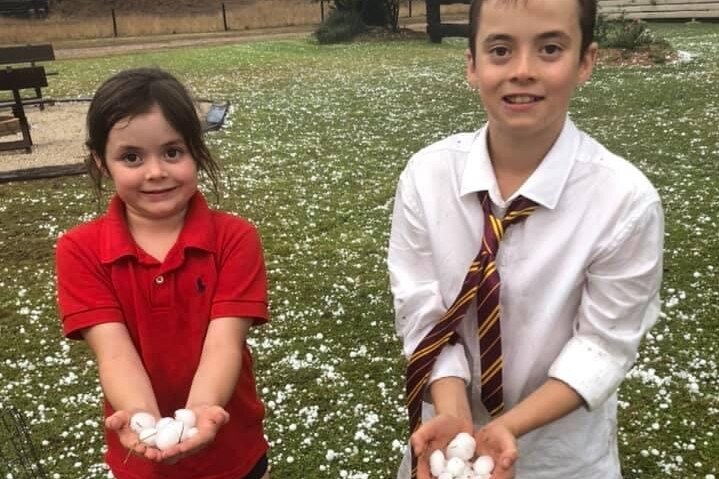 A young girl and boy hold hail stones in their outreached hands smiling at the camera. The ground littered in hail stones