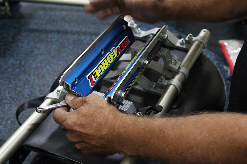 A photo of a man using tools to join two blue, red and yellow blades to the underside of a black plastic ice sledge hockey sled.