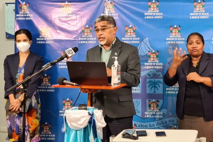 Wide shot of a man speaking at a lectern, flanked by two women.