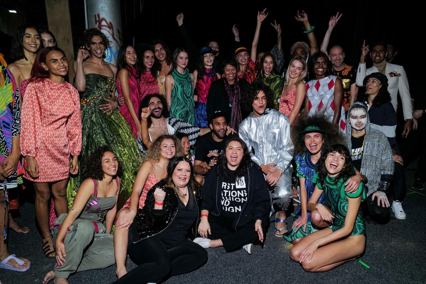 Grace Lillian Lee cheers, seated on the ground, surrounded by Indigenous models and designers