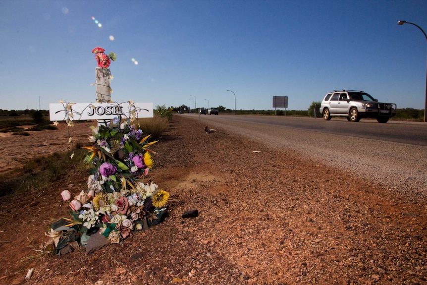 A makeshift crucifix, emblazoned with "josh" stands, wreathed by flowers, by the side of a dusty red road.