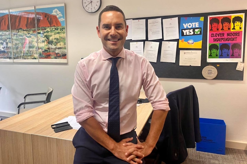 a young man smiling while sitting on his desk in an office