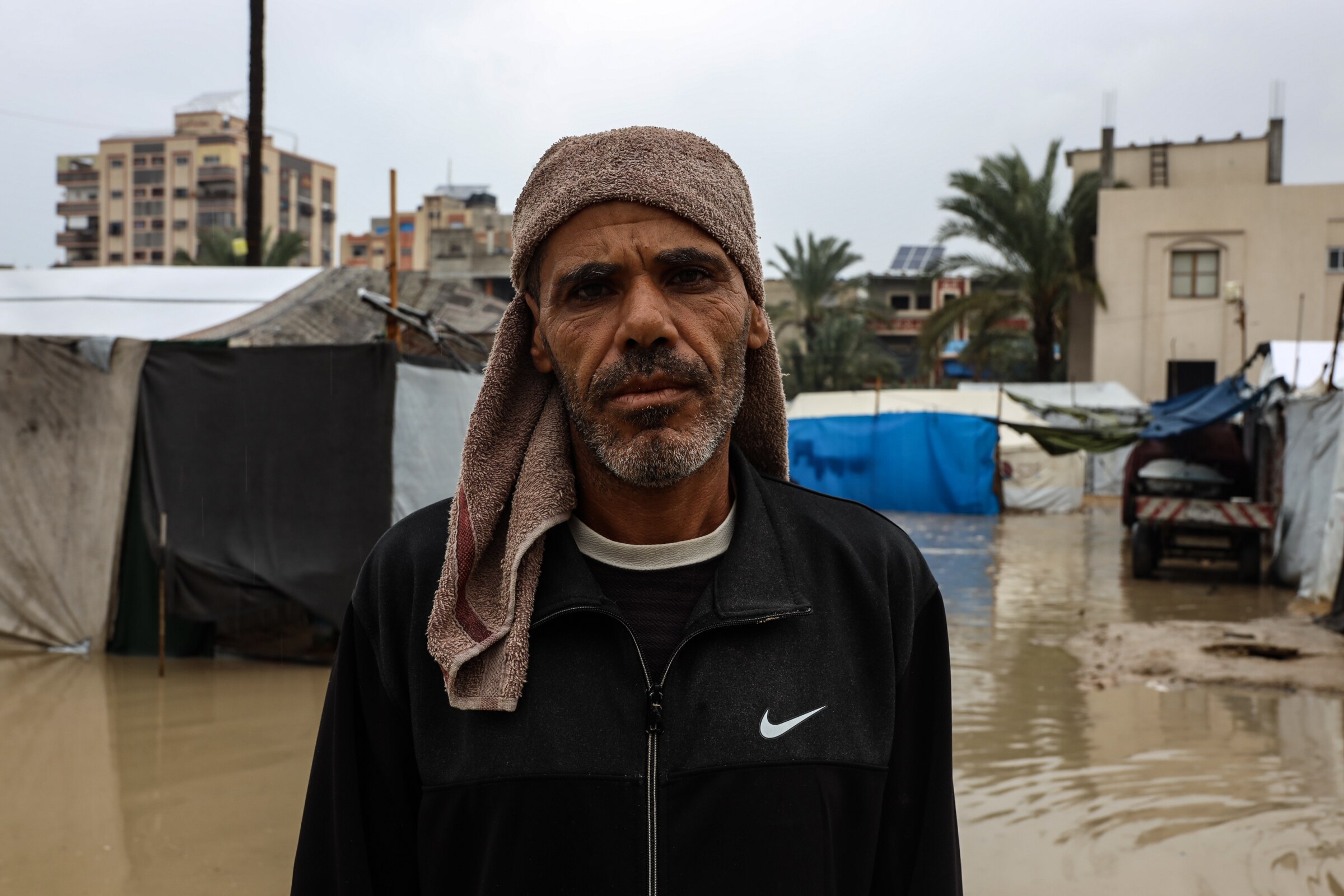 A man with a towel wrapped around his head stands in front of tents which are in muddy water.