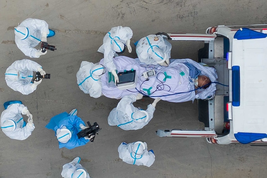 A photo taken from directly above shows a patient being loaded into an ambulance with medics wearing protective suits nearby.