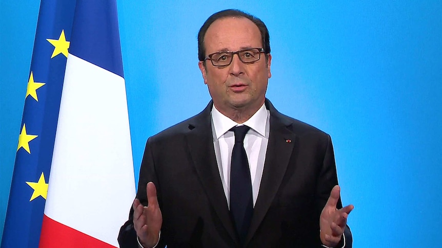 French president looking straight ahead, hands up, his mouth slightly open to talk and a flag in the background