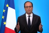 French president looking straight ahead, hands up, his mouth slightly open to talk and a flag in the background