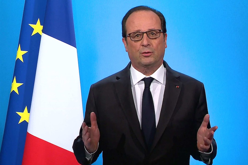 French president looking straight ahead, hands up, his mouth slightly open to talk and a flag in the background