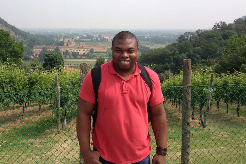 Edward Obi stands in front of a winery wearing a red tshirt and black backpack straps