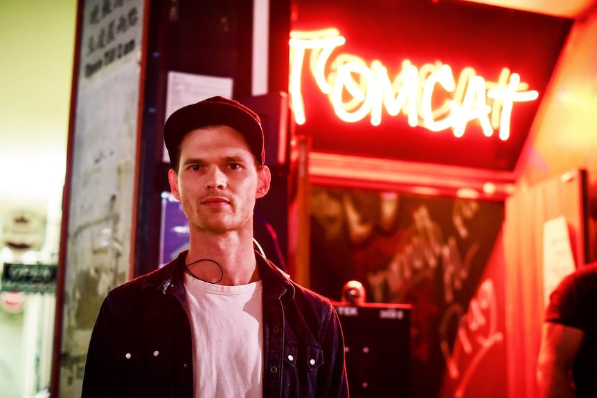 A man in cap stands in front of a red fluro sign outside the nightclub tomcat