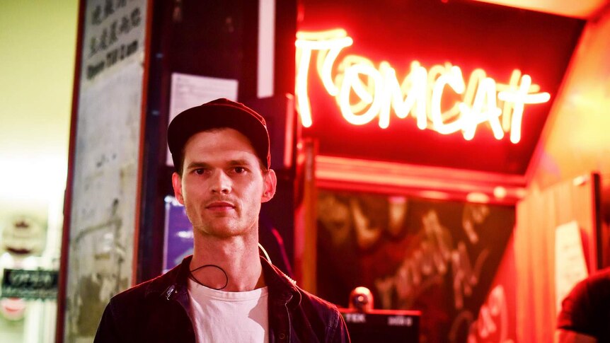 A man in cap stands in front of a red fluro sign outside the nightclub tomcat