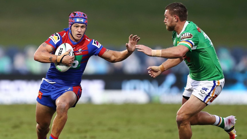 A Newcastle Knights NRL players holds the ball in his right hand and he uses his left to fend off an opponent.
