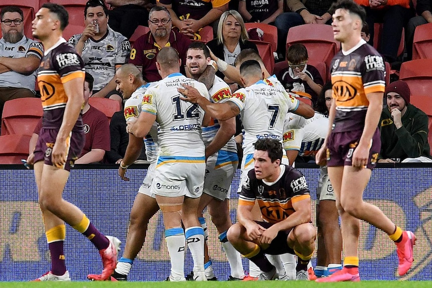 Gold Coast NRL players embrace as they celebrate a try as Broncos players walk past in the foreground.