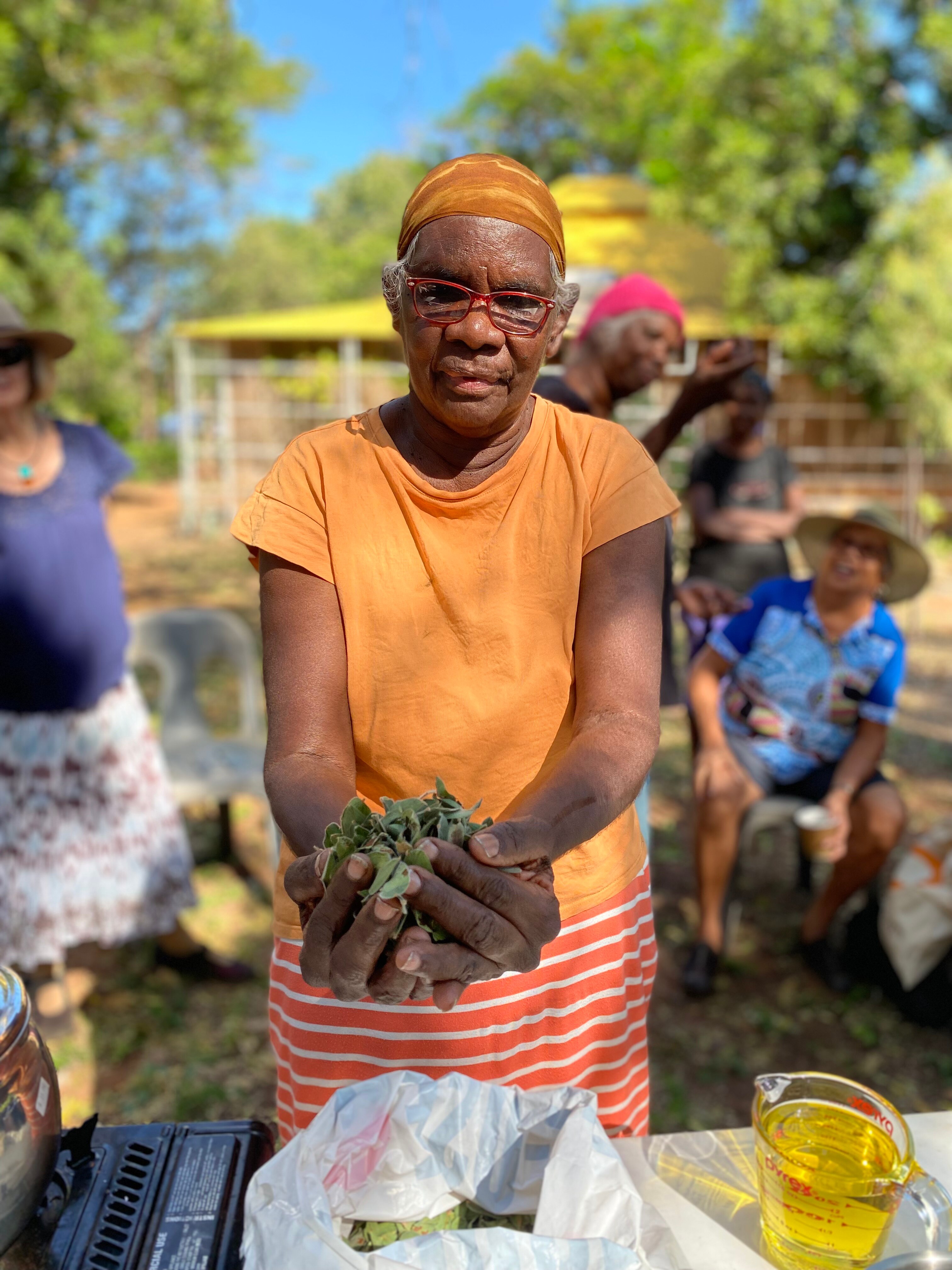 An Indigenous woman with an orange bandana and shirt holds a bunch of cockroach bush