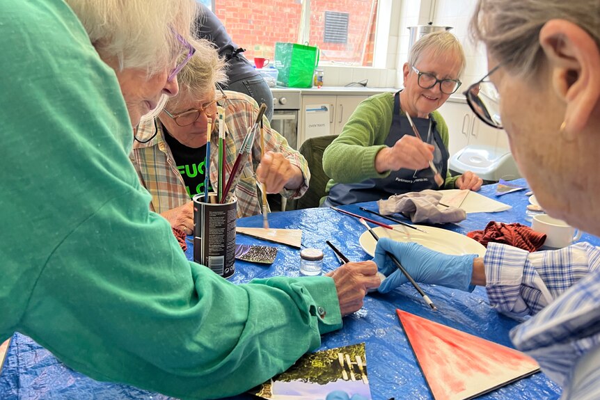 Older woman in green overcoat leans over shared art table surrounded by other other older female artists