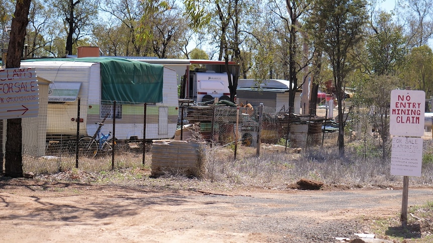 'No entry mining claim', 'for sale' signs, trees, dirt road and temporary structures, caravans in background.