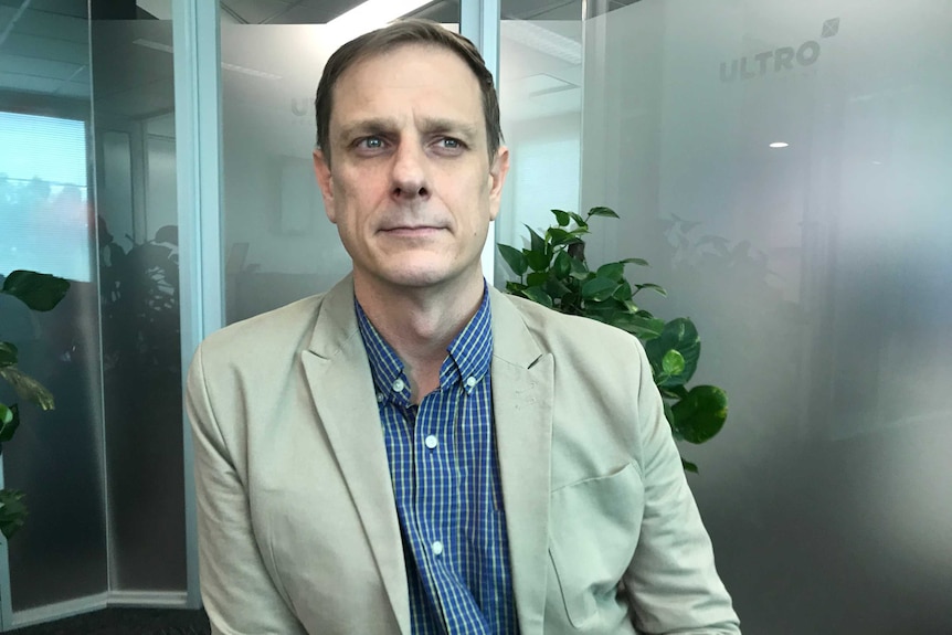 Image of a man standing in an office wearing a cream coloured blazer and blue checked shirt.