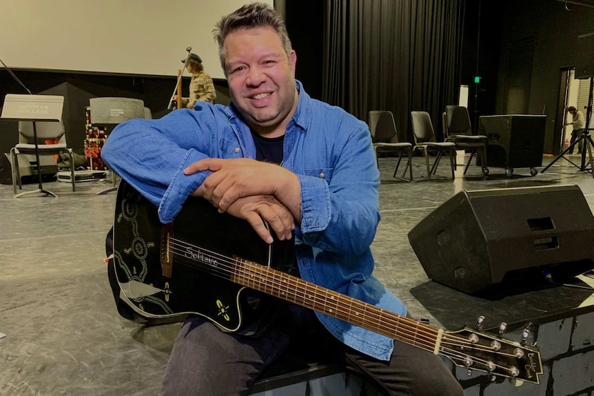 Phil Walleystack sits on the edge of a stage holding a guitar.