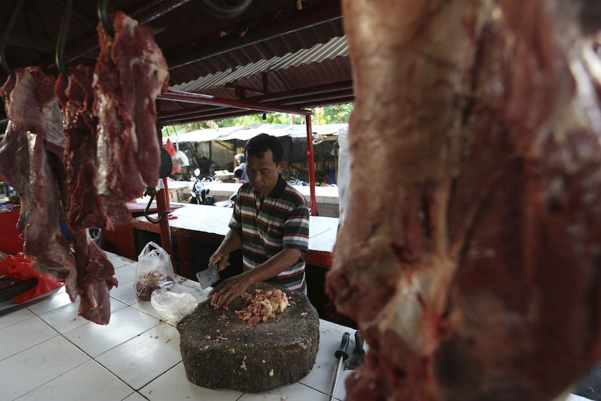 A man carves up meat with a cleaver on a butcher's block at a stall lined with hanging meat at markets.