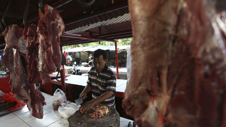 A man carves up meat with a cleaver on a butcher's block at a stall lined with hanging meat at markets.