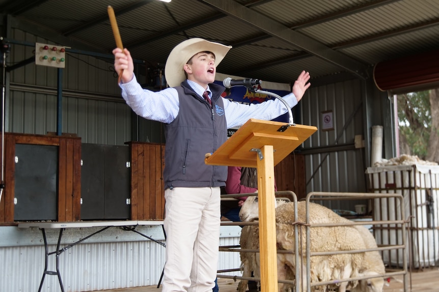 A teenage boy in a hat, vest and business shirt gestures while he conducts an auction.