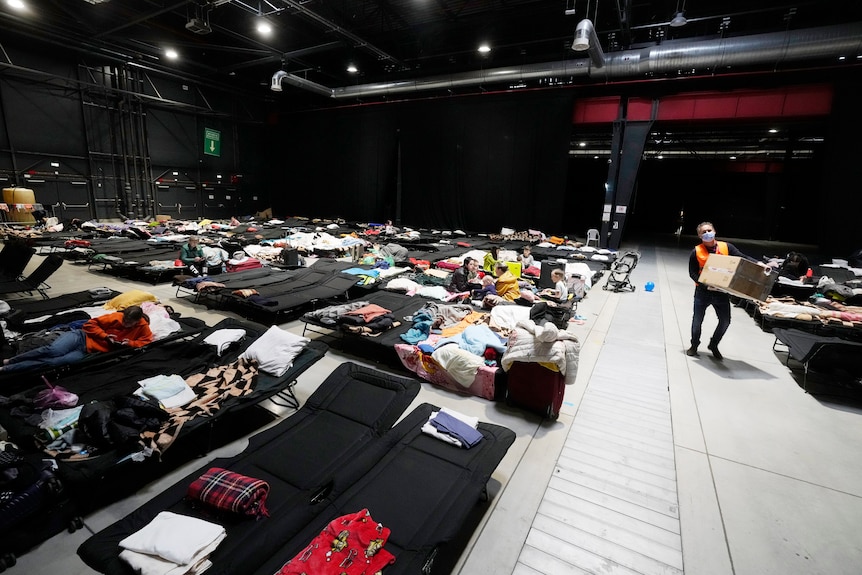 A large hall, in which mattresses and lounge chairs are set up, and some people lie on them. A man walks by with a box.