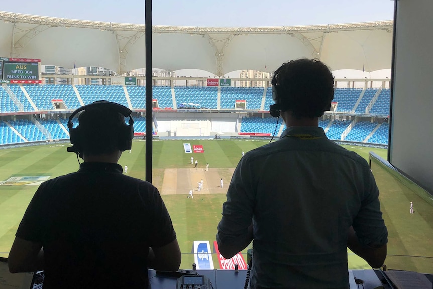 Wide shot from behind of two men standing looking out a window onto a cricket ground.