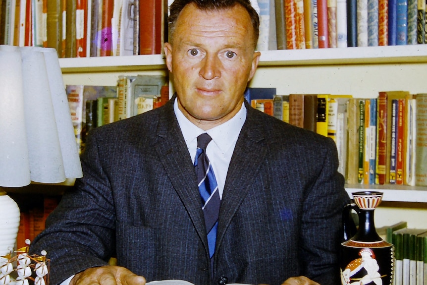 A middle-aged man in a suit and tie in front of a bookcase.