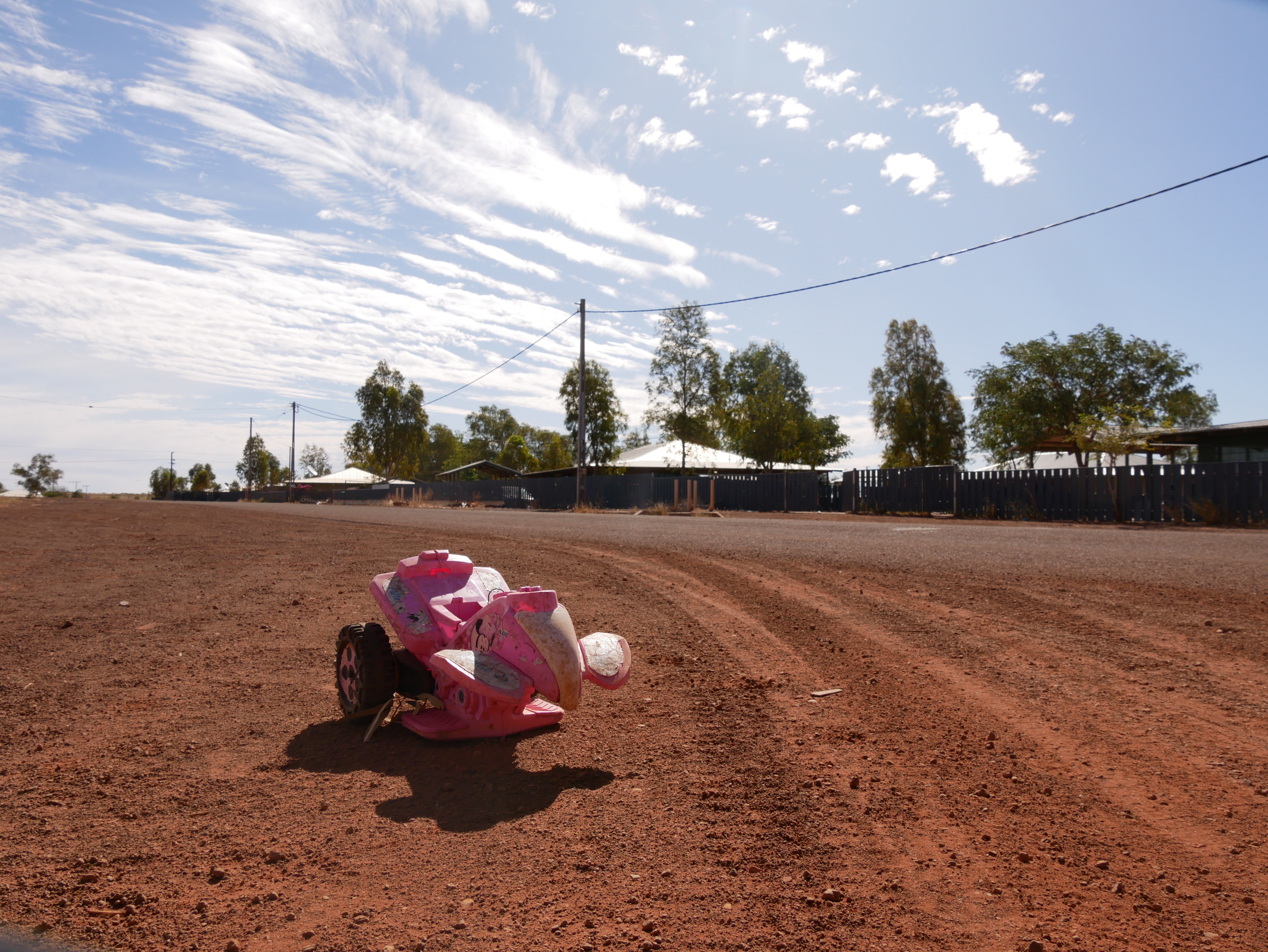 A wide red dirt road with houses in background and children's toy in foreground