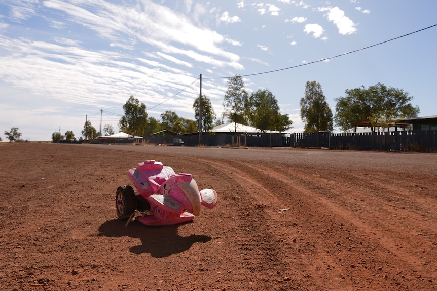 A wide red dirt road with houses in background and children's toy in foreground.