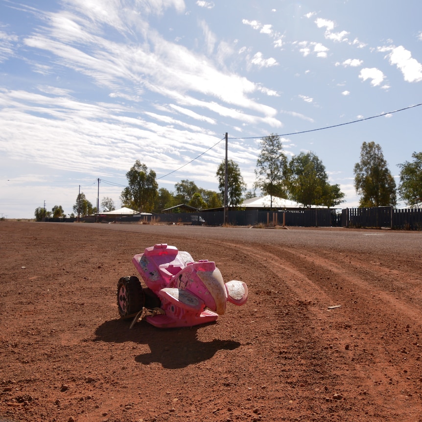 A wide red dirt road with houses in background and children's toy in foreground