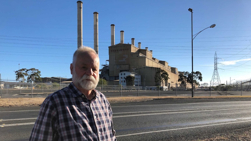 An older man with a beard stands in front of a power station.