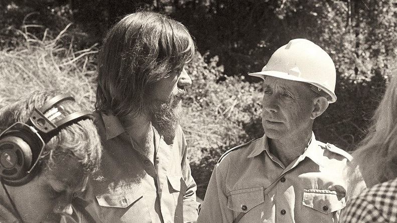 A black and white photo of a man in a helmet talking to a man with a beard. Journalists stand nearby.