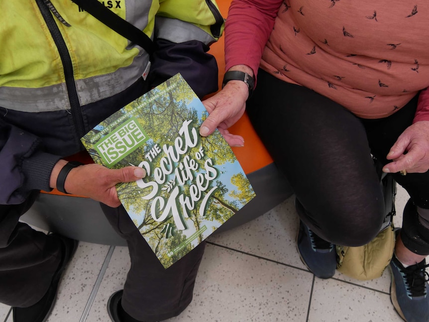 A man and a woman holding an issue of The Big Issue while sitting down on a bench.