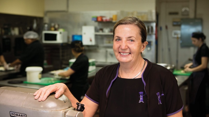 A woman in a black chef's uniform stands in a kitchen, smiling.