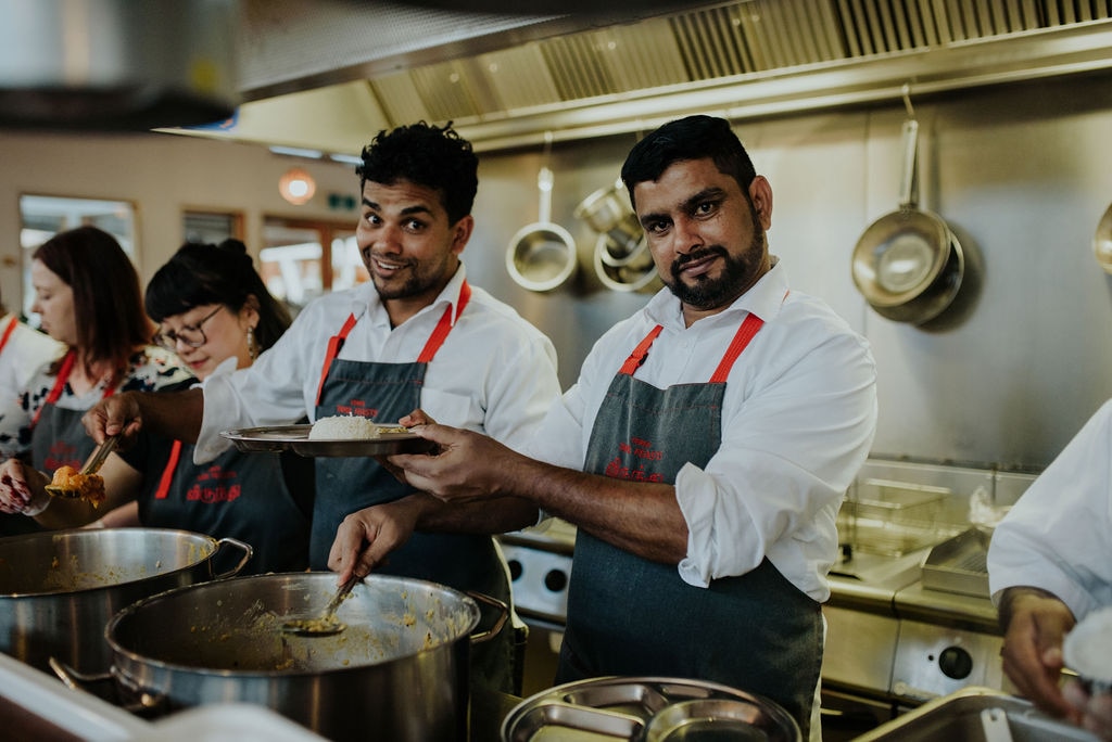 Three people wearing aprons and white shirts stand behind large, steaming pots, smiling widely.