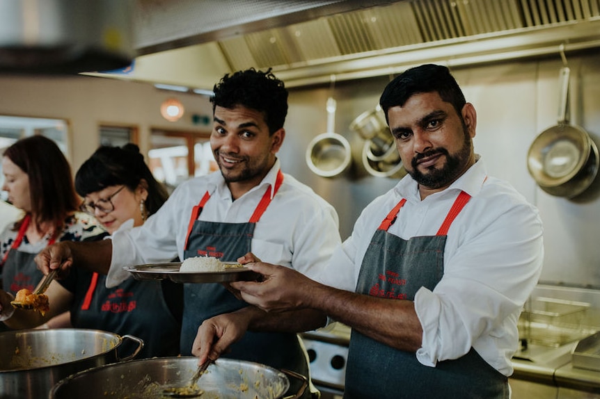 Three people wearing aprons and white shirts stand behind large, steaming pots, smiling widely.