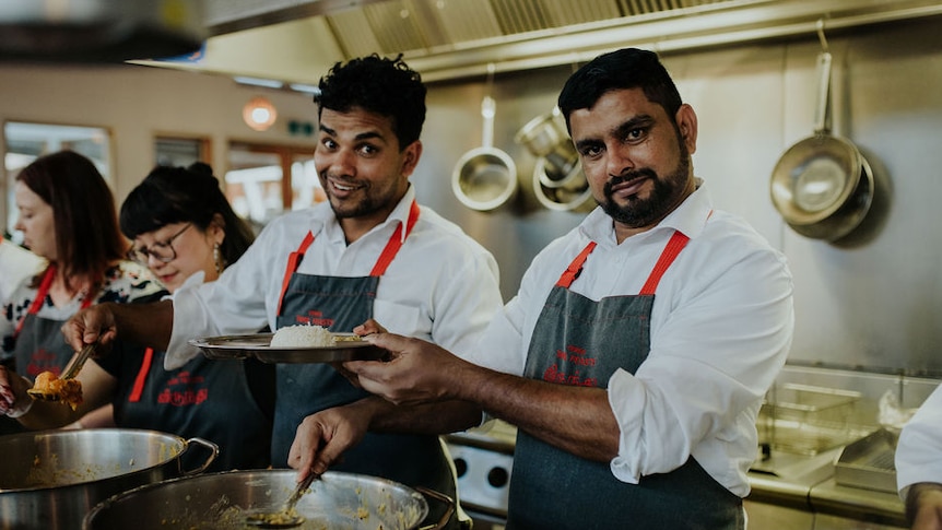 Three people wearing aprons and white shirts stand behind large, steaming pots, smiling widely.