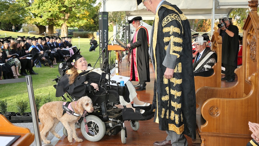 A woman in a wheelchair with a dog wears a graduation gown and hat. She is near two people standing in black robes.